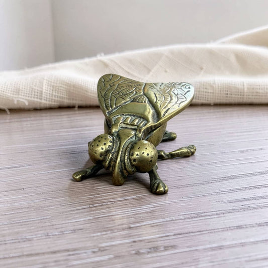 A brass fly-shaped door dish resting on a light wood surface with a beige fabric background.