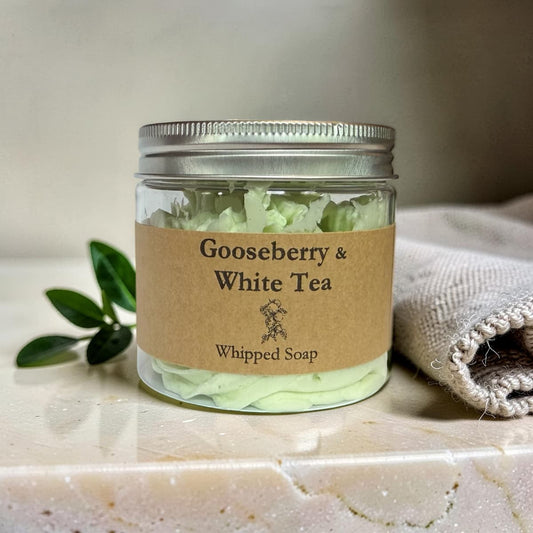 A clear glass jar of Whipped Soap 'Gooseberry & White Tea' with a kraft paper label and silver metal lid, set on a marble surface next to a green sprig and a beige cloth.
