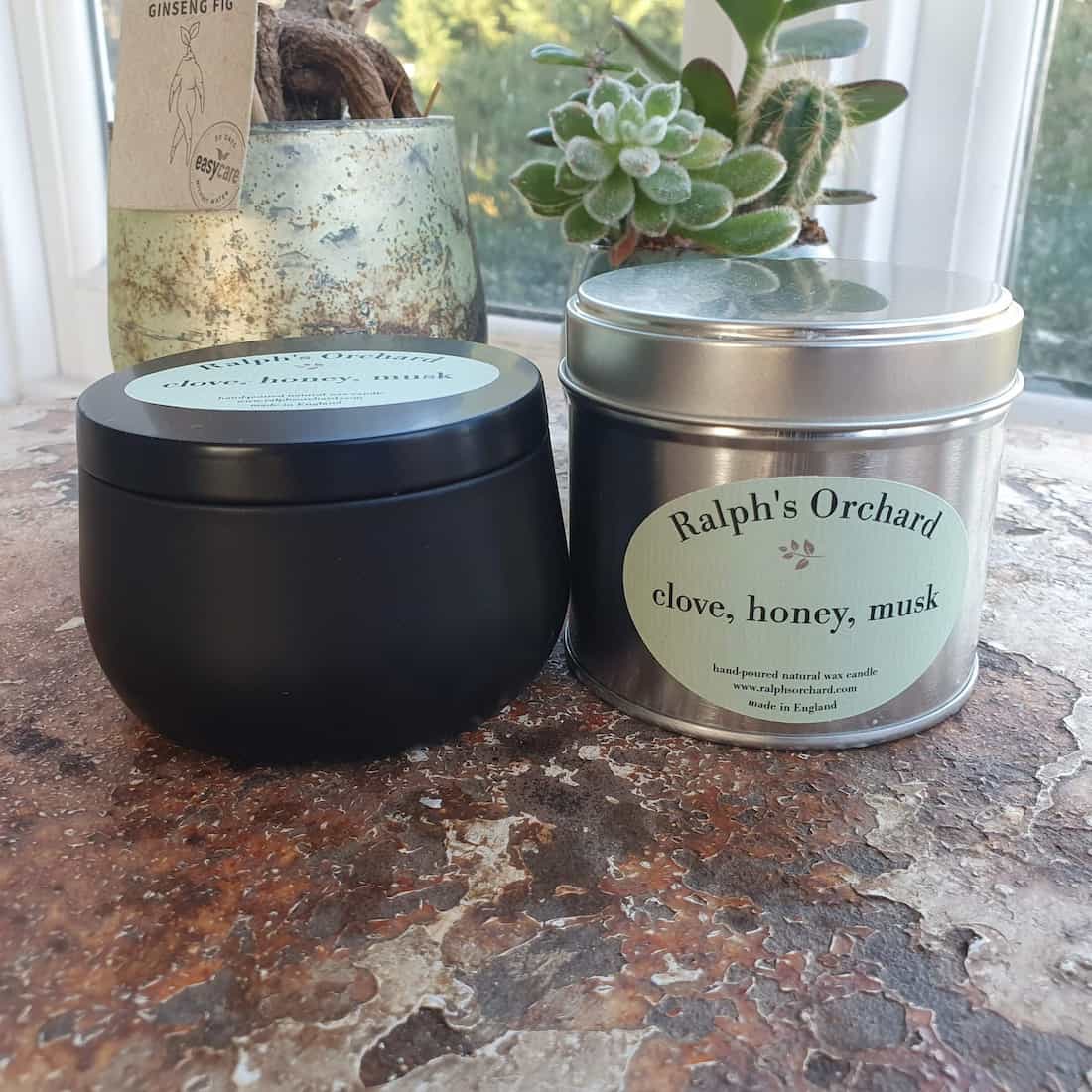 Two candles from Ralph's Orchard on a textured surface with plants in the background