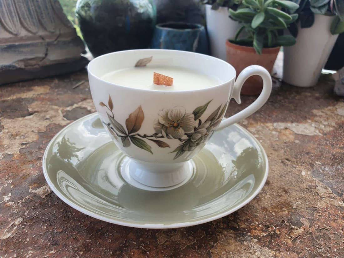 Teacup with floral design containing a candle on a saucer, with plants in the background.