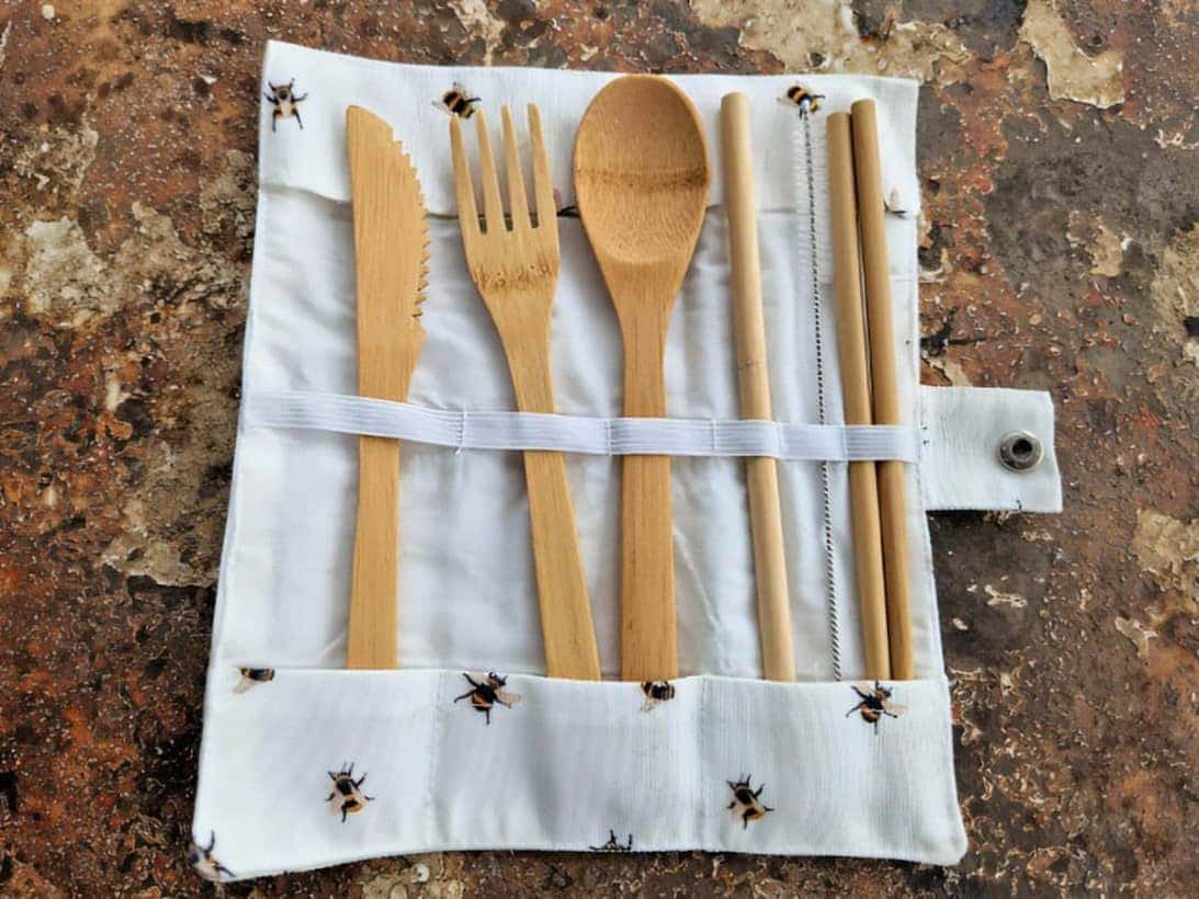 Set of bamboo cutlery including knife, fork, spoon, and straws on a white roll-up case with bee pattern against a rustic background.