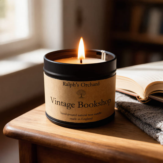 A lit candle in a black metal tin with a tan paper label reading 'Ralph's Orchard Vintage Bookshop,' placed on a wooden table beside an open book and a folded gray knit blanket in a softly lit indoor setting.