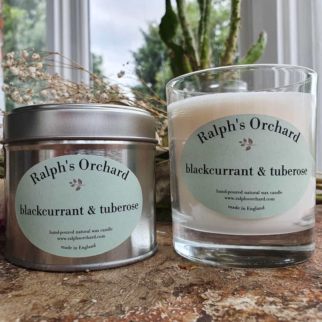Two candles labeled 'Ralph's Orchard' on a wooden surface with a blurred natural background.