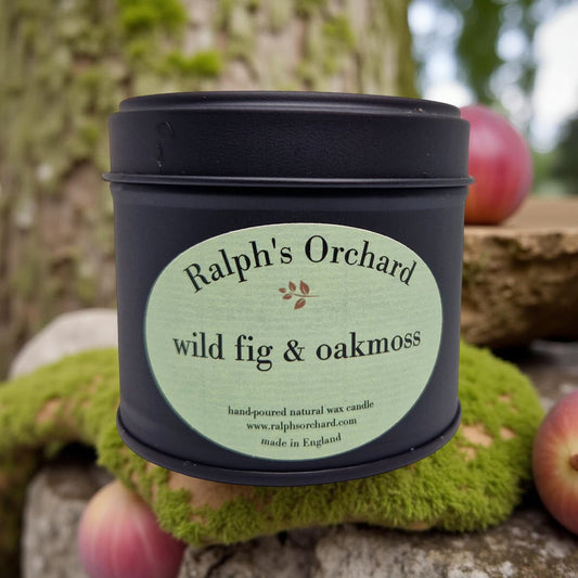 Candle in a black tin labeled 'Ralph's Orchard wild fig & oakmoss' against a natural background.