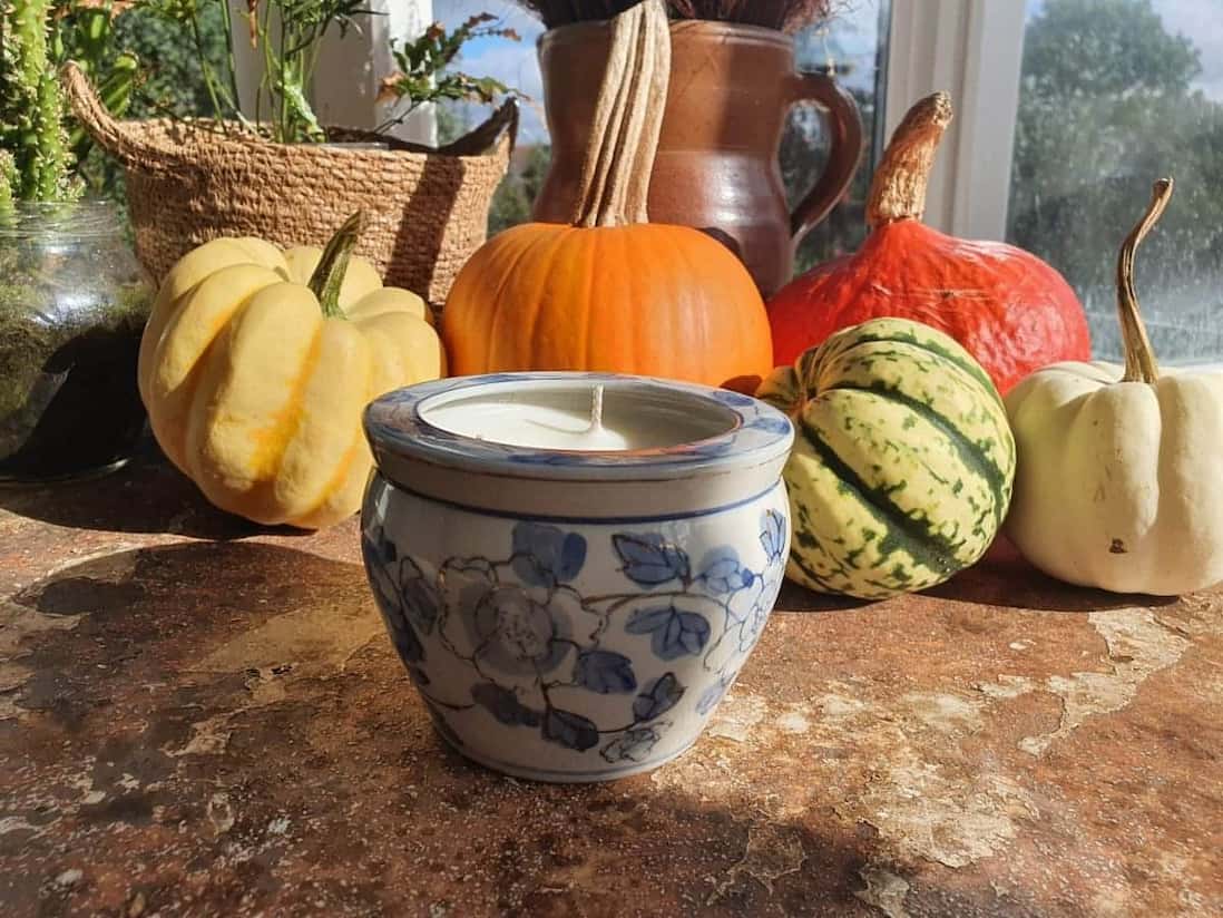 Ceramic container with a lit candle surrounded by pumpkins and gourds on a windowsill.