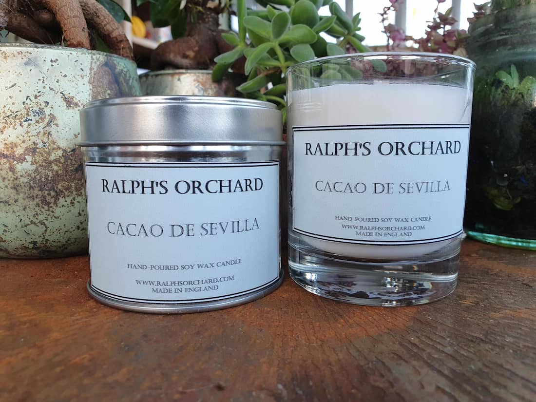 Two candles from Ralph's Orchard on a wooden surface with plants in the background