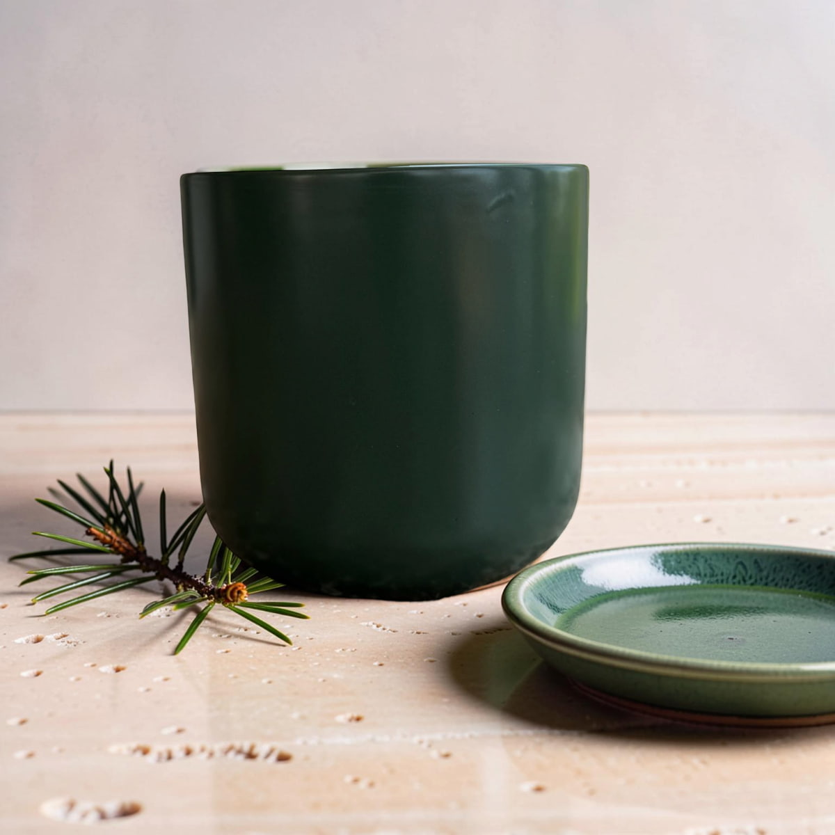 A dark green ceramic planter pot with a matching small saucer, styled on a light beige stone surface with a pine sprig.