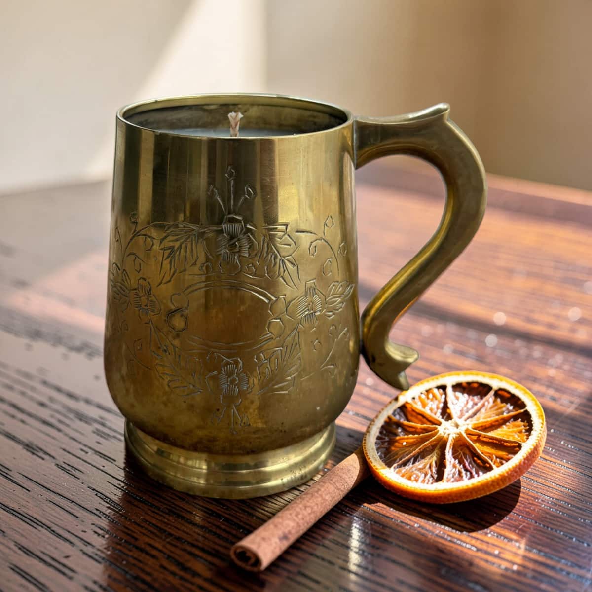 A brass mug candle with floral engravings sits on a dark wooden table next to a dried orange slice and a cinnamon stick.