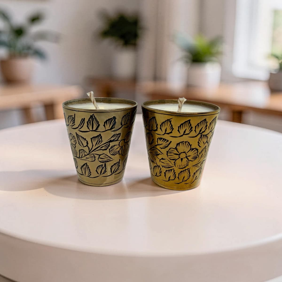 Two decorative metal candle holders with floral engravings, each containing a white candle, placed on a round white table in a softly blurred indoor setting.