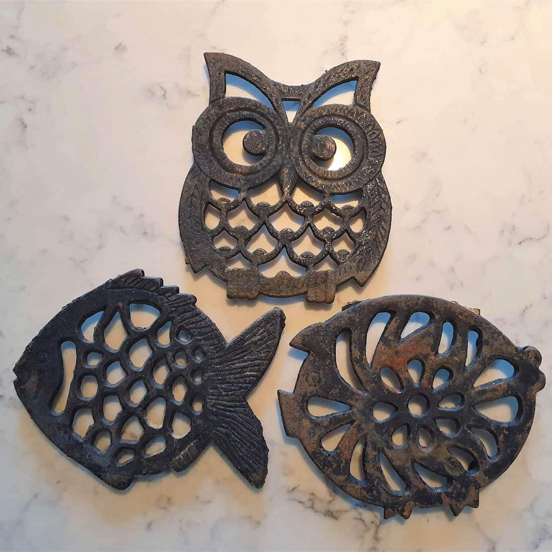 Three cast iron trivets shaped like an owl, fish, and flower on a marble surface.