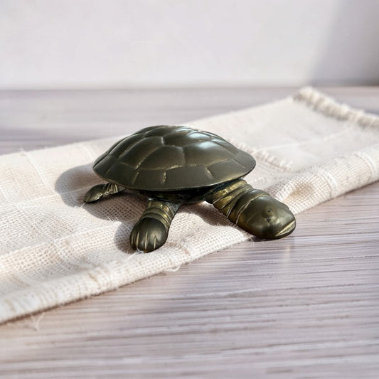 A small metallic turtle figurine with a dark bronze finish, resting on a cream-colored woven textile over a light wood surface.