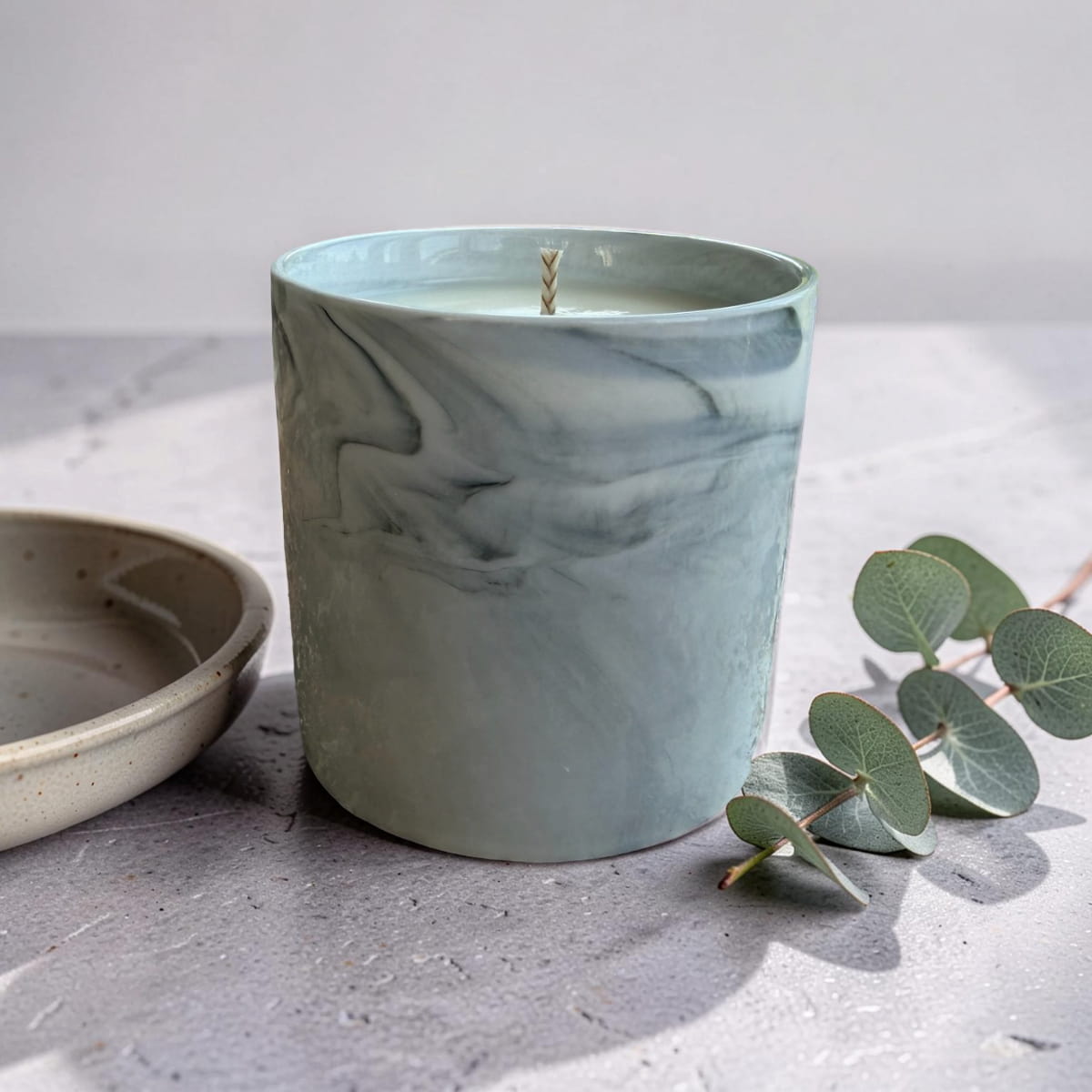 A marble-patterned teal candle with a single wick sits on a gray stone surface next to a ceramic dish and eucalyptus leaves.