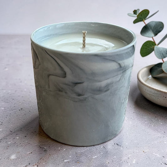A marble-patterned ceramic candle jar with a light sage green and gray swirl design, filled with white wax and featuring a single wick, set on a textured gray surface next to a small eucalyptus sprig.