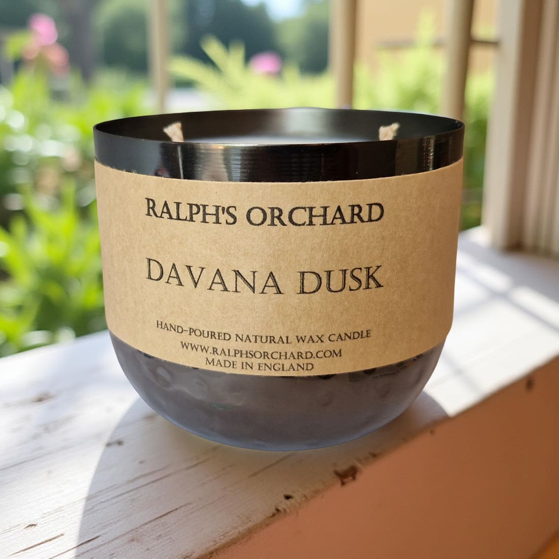 Candle labeled 'Ralph's Orchard Davana Dusk' on a wooden surface with a blurred green outdoor background.
