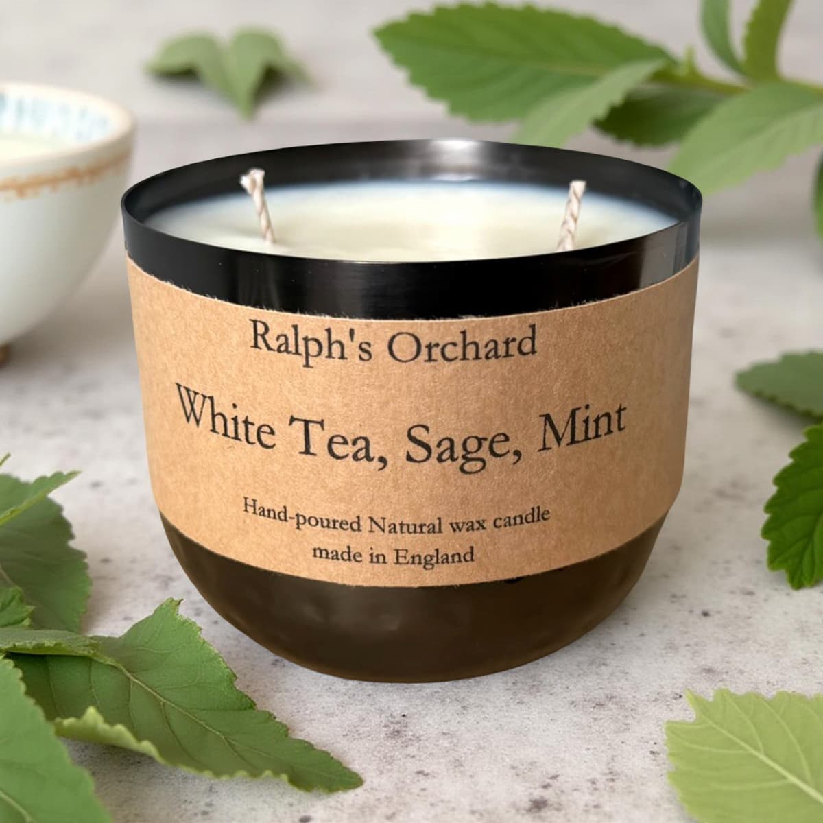 Candle labeled 'Ralph's Orchard White Tea, Sage, Mint' on a surface with green leaves.