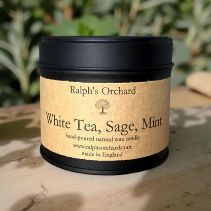 Candle in a tin labeled 'White Tea, Sage, Mint' by Ralph's Orchard on a natural surface with greenery in the background.