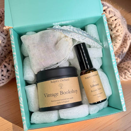 A gift set from Ralph's Orchard in a mint green box, featuring a jar of 'Vintage Bookshop' natural wax candle and a bottle of 'Library Romance Mist', surrounded by white packing foam, a white drawstring bag, and a silver feather ornament.