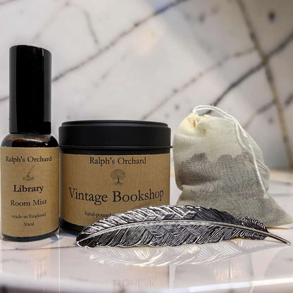 Ralph's Orchard 'Vintage Bookshop' hand-poured room mist and matching jar displayed on a reflective white surface with a feather and drawstring pouch in front, against a marble-patterned background.