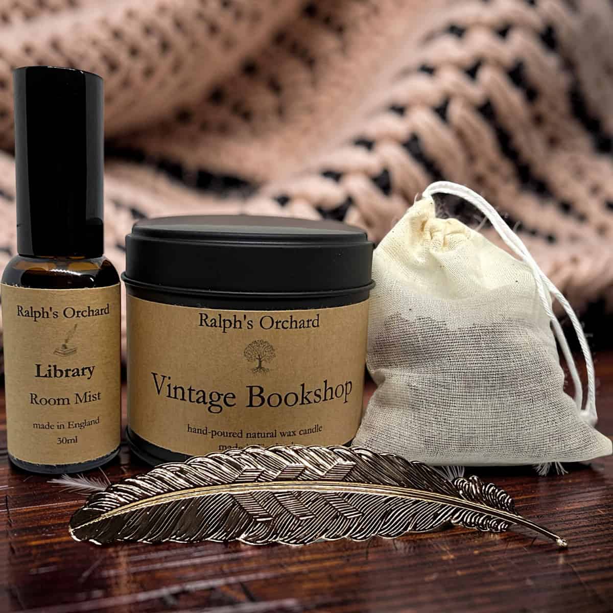 A gift set from Ralph's Orchard featuring a 'Vintage Bookshop' candle jar, a 'Library Room Mist' spray bottle, and a small cream drawstring pouch, styled with a feather on a wooden surface.