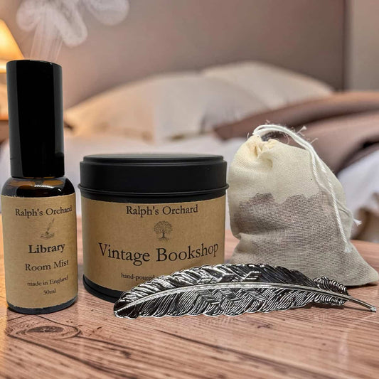 A styled product arrangement on a wooden surface featuring a small amber bottle of Ralph's Orchard Library Room Mist with a black cap, a matching black tin of Ralph's Orchard Vintage Bookshop hand towels, a beige drawstring fabric pouch, and a decorative silver feather quill on a wood tabletop with a softly blurred bedroom background.