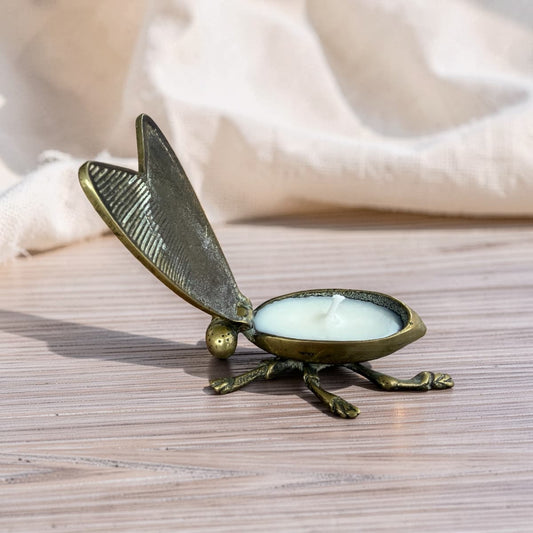 An antique-style brass insect candle holder shaped like a beetle, holding a white tea light candle, set on a light wood surface with a soft beige fabric background.