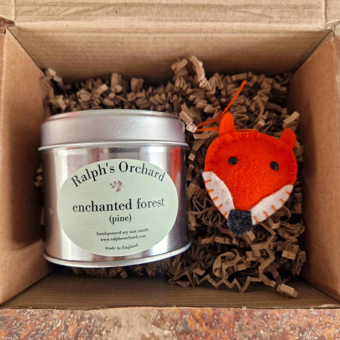 Candle labeled 'Ralph's Orchard' with a felt fox ornament in a cardboard box.