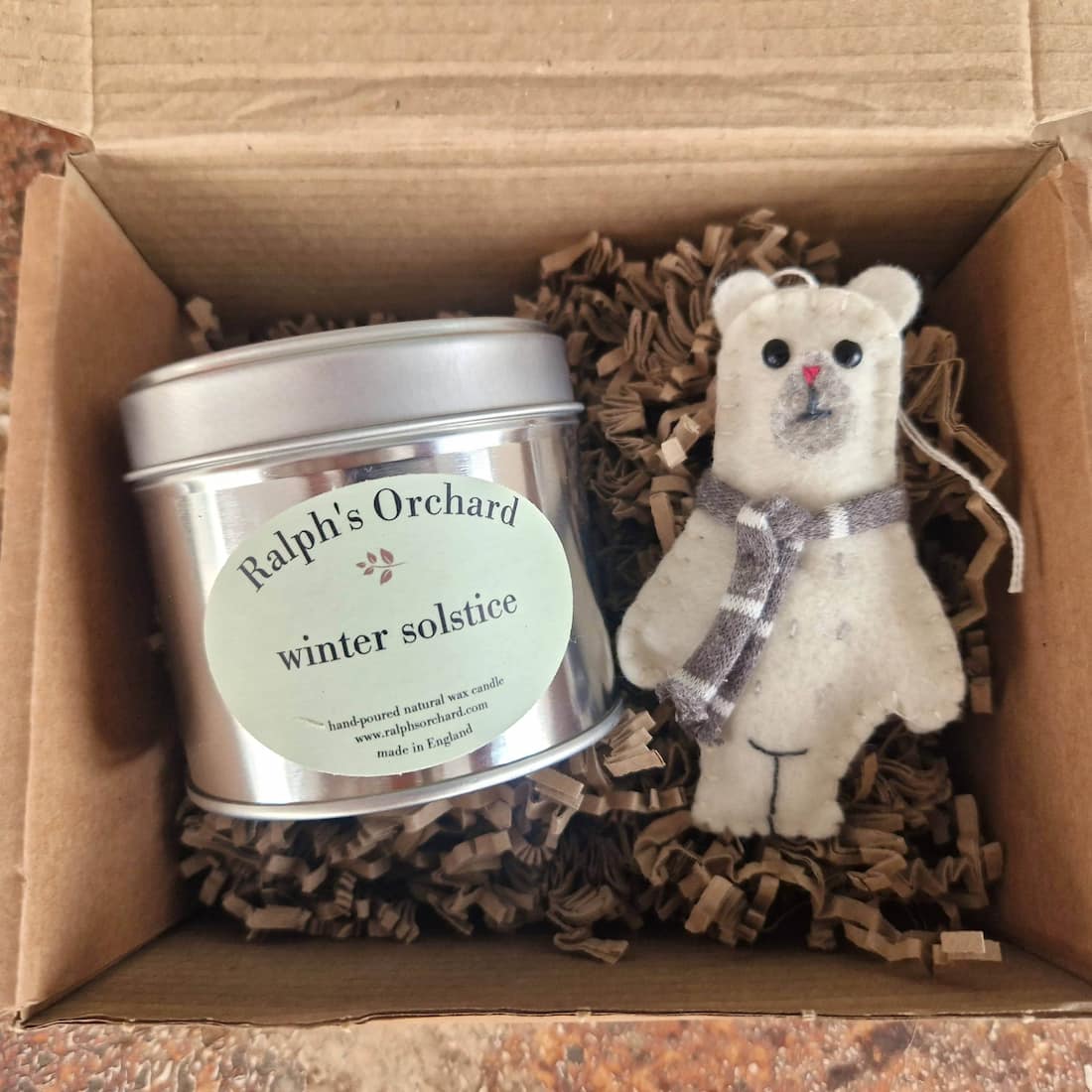 Candle labeled 'Ralph's Orchard winter solstice' and a small teddy bear with a bow in a cardboard box.