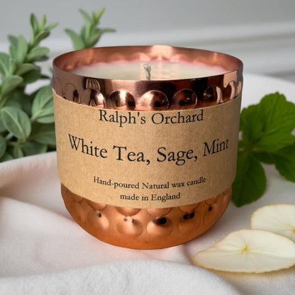 Candle with a copper holder and 'Ralph's Orchard' label on a white surface with greenery.