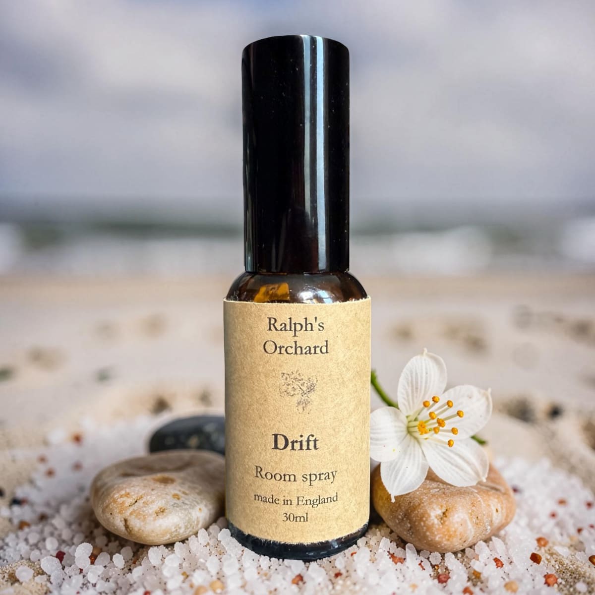 Bottle of Ralph's Orchard Drift room spray on a pebble surface with a white flower.
