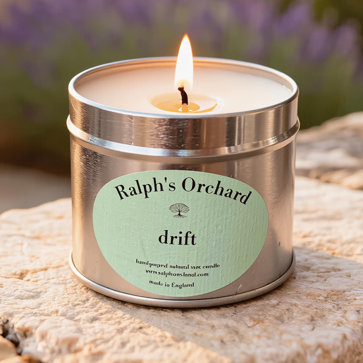 Candle in a metal tin labeled 'Ralph's Orchard drift' on a stone surface with blurred lavender in the background.