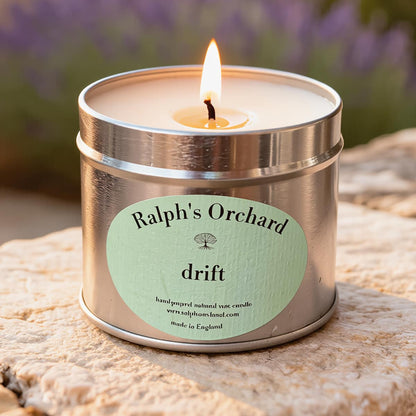 Candle in a metal tin labeled 'Ralph's Orchard drift' on a stone surface with blurred lavender in the background.
