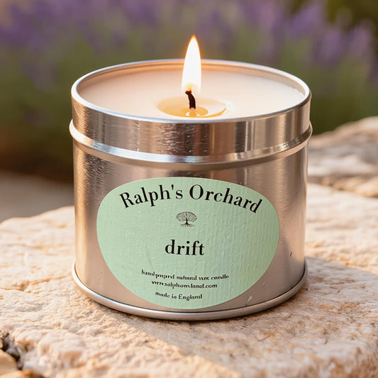 Candle in a metal tin labeled 'Ralph's Orchard drift' on a stone surface with blurred lavender in the background.