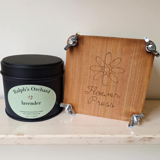 Black candle labeled 'Ralph's Orchard lavender' next to a wooden flower press on a light surface.