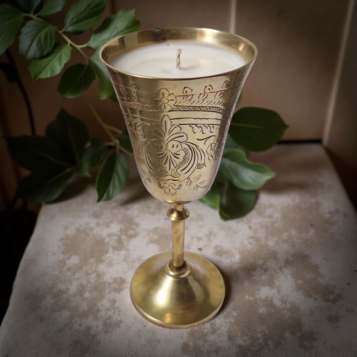 Decorative brass candle holder with a lit candle on a textured surface with green leaves in the background