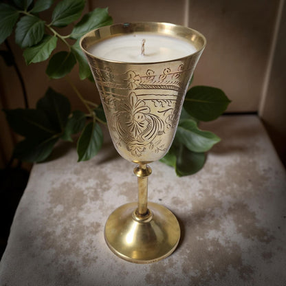 Decorative brass candle holder with a lit candle on a textured surface with green leaves in the background