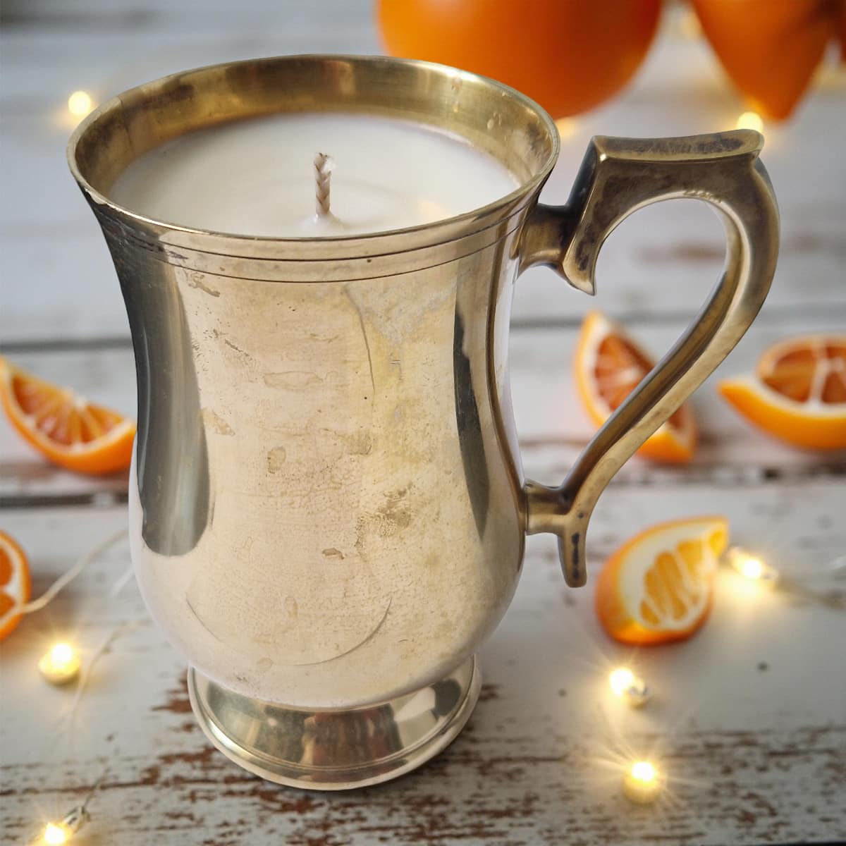 Candle in a metallic mug with oranges and lights on a wooden surface