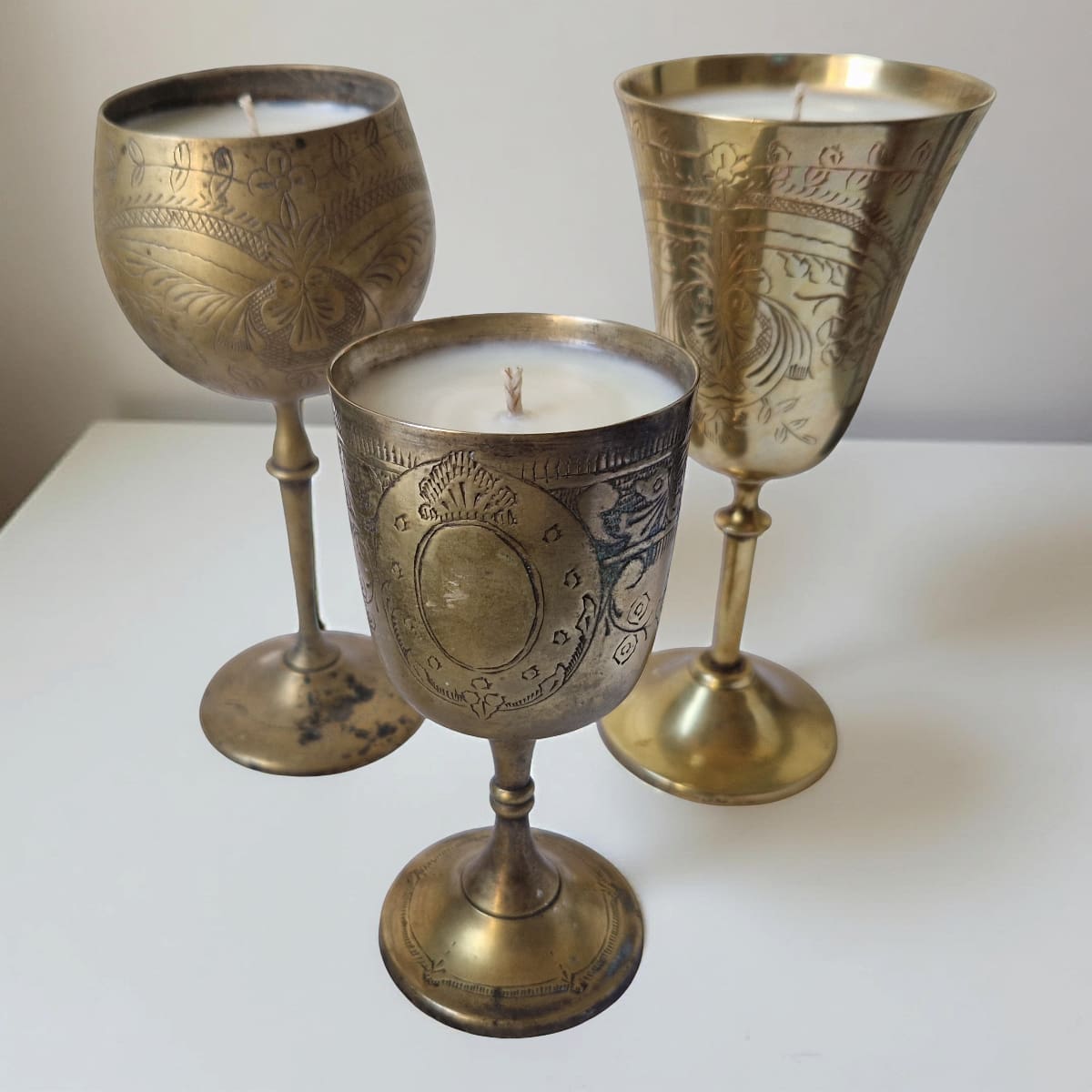 Three ornate brass candle holders with candles inside on a white surface.