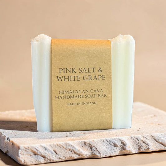A handmade soap bar wrapped in a kraft paper label reading 'PINK SALT & WHITE GRAPE', set on a stone slab against a warm beige background.