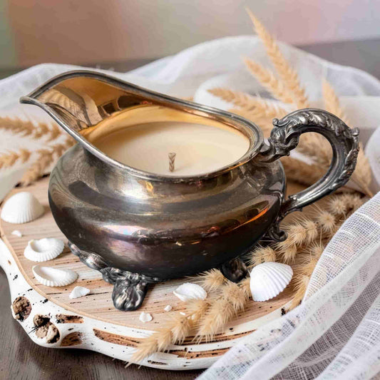 Candle in a silver gravy boat with seashells and dried grasses on a wooden board.