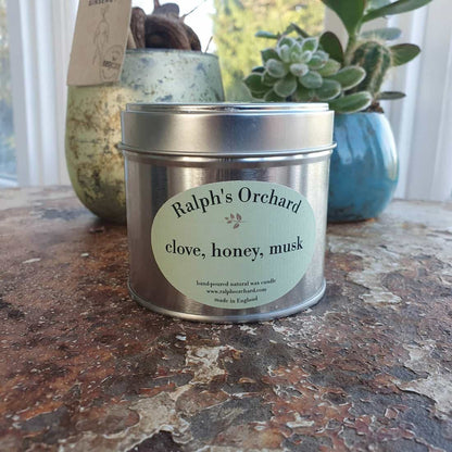 Candle tin labeled 'Ralph's Orchard' with a plant in the background