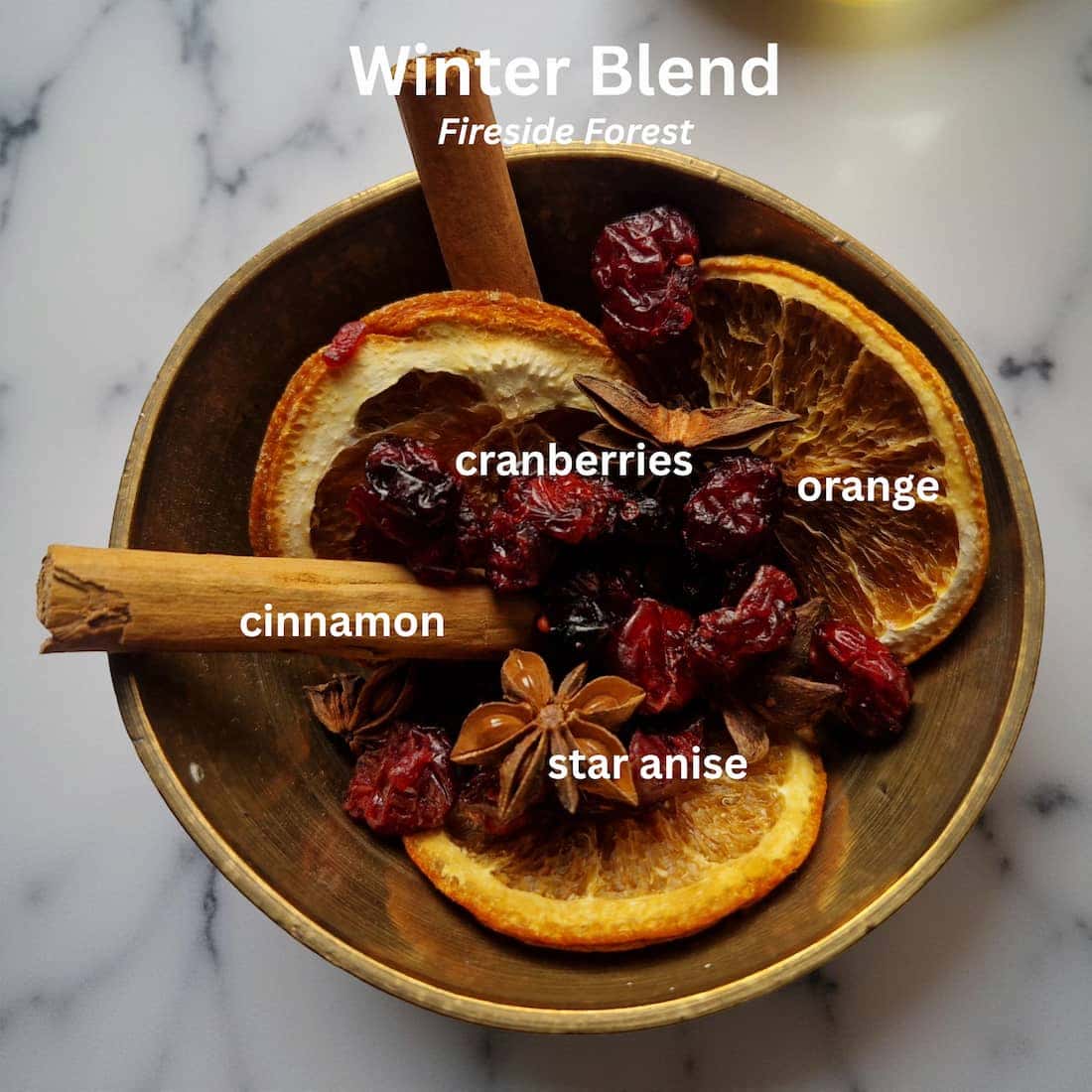 Wooden bowl with winter blend ingredients including oranges, cranberries, cinnamon, and star anise on a marble surface.