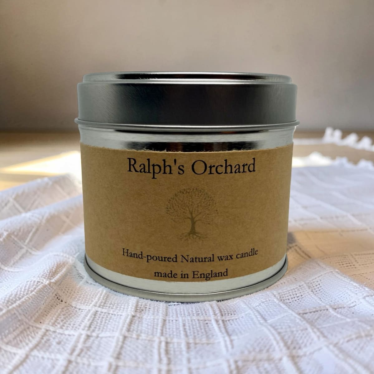 A silver metal tin candle with a brown kraft paper label reading 'Ralph's Orchard' and 'Hand-poured Natural wax candle made in England,' set on a white textured cloth.