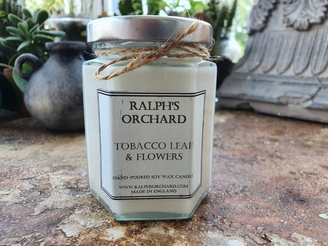 Candle jar labeled 'Ralph's Orchard Tobacco Leaf & Flowers' on a stone surface with plants in the background.