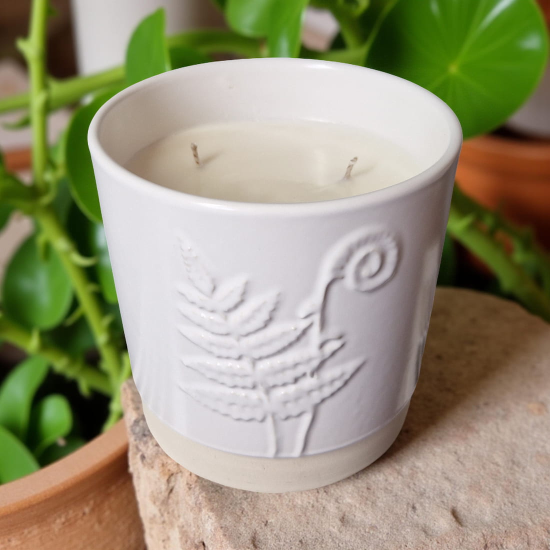 White ceramic candle with embossed design on a stone surface with greenery in the background