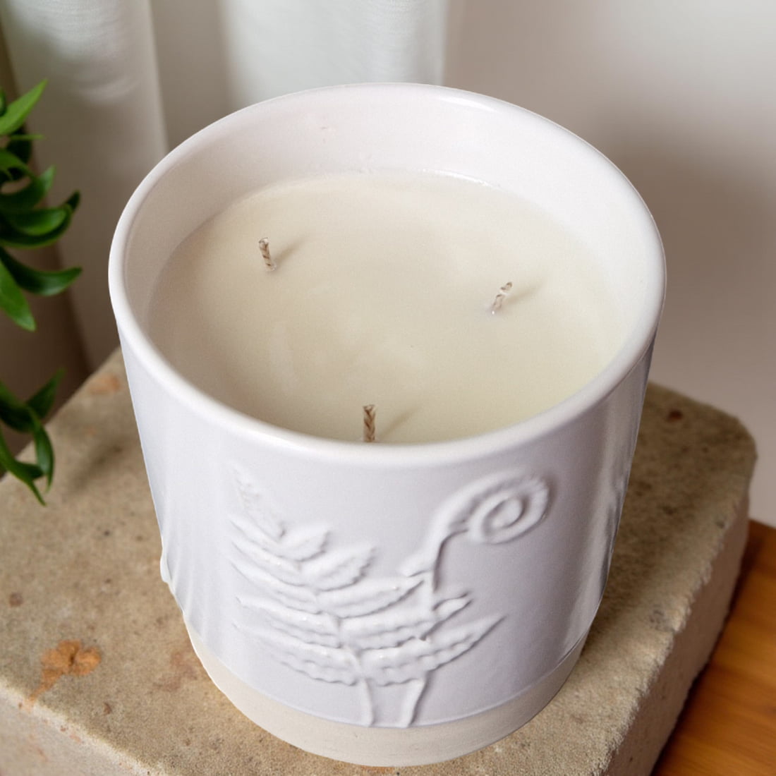 White candle in a decorative container on a stone surface with a plant in the background