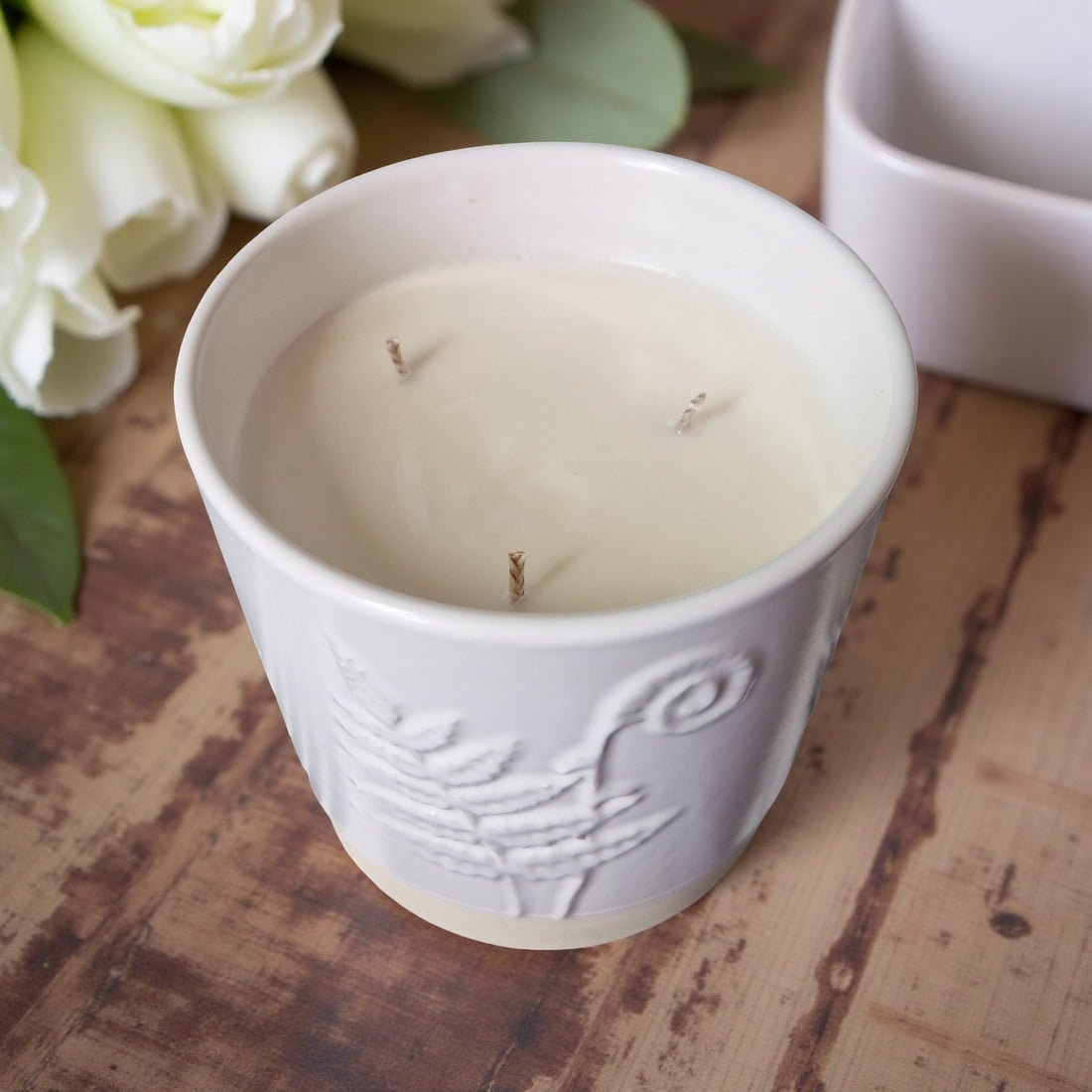 White candle in a decorative container on a wooden surface with white flowers in the background