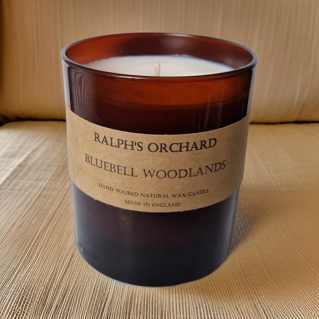 Candle with a label reading 'Ralph's Orchard Bluebell Woodlands' on a woven surface.