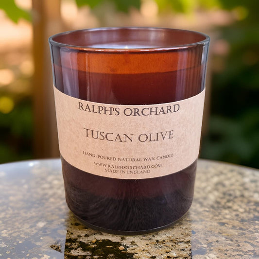 Candle labeled 'Ralph's Orchard Tuscan Olive' on a reflective surface with a blurred natural background