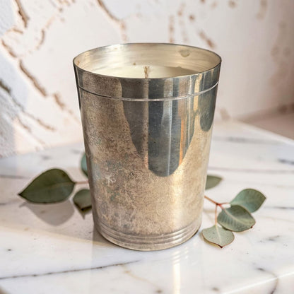 Silver metallic candle holder with a candle on a marble surface with eucalyptus leaves.