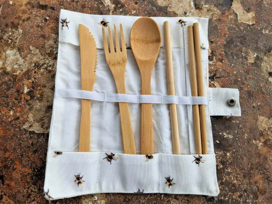 Set of bamboo cutlery including knife, fork, spoon, and straws on a white roll-up case with bee pattern against a rustic background.